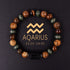 Natural zodiac stone bracelet showing three-color alternating beads — warm chatoyant tiger eye, blue-green hawk eye and dark agate — with engraved black obsidian zodiac symbol charm on elastic cord.