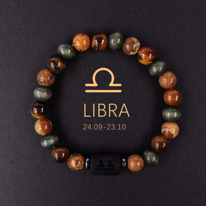 Natural zodiac stone bracelet resting on dark wood beside a leather watch and linen cloth, soft daylight, masculine grounded zodiac atmosphere, handmade crystal jewelry.